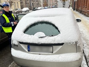 Zdjęcie przedstawia nieodśnieżony samochód i policjanta